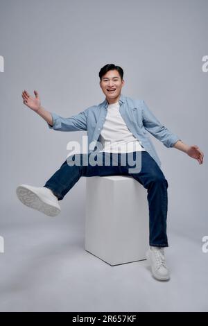 Lächelnder, glücklicher asiatischer Mann, der im Studio auf dem Würfel sitzt, mit weißem Hintergrund Stockfoto