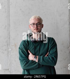 Ein Mann mit blauen Haaren, einer Brille und einem grünen Sweatshirt posiert selbstbewusst mit gekreuzten Armen vor grauem Hintergrund und zeigt sein modisches Gesicht Stockfoto