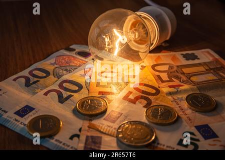 Glühbirne leuchtet, mit Münzen und Banknoten daneben. Steigende Stromtarife, Energieabhängigkeit, Energiequellen und Energieversorgung. Stockfoto