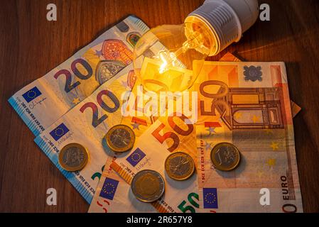 Glühbirne leuchtet, mit Münzen und Banknoten daneben. Steigende Stromtarife, Energieabhängigkeit, Energiequellen und Energieversorgung. Stockfoto