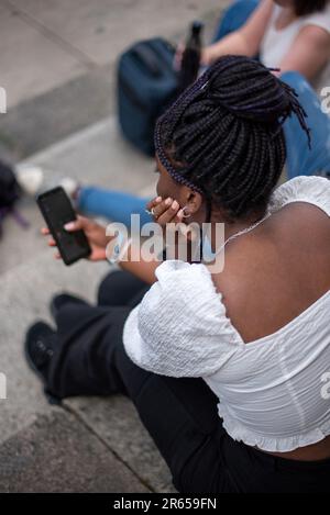 Ein schwarzes Teenager-Mädchen mit Zöpfen in einem weißen Oberteil sitzt in Gesellschaft von Freunden auf der Straße und schaut auf das Telefon. Rückansicht. Stockfoto