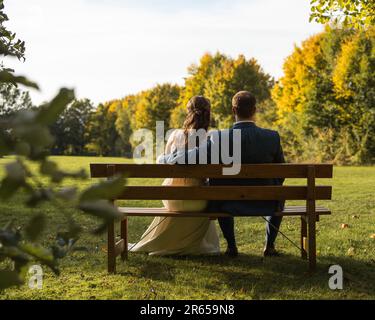 Ein Hochzeitsfoto eines Paares, das einen sonnigen Tag in einem Park genießt, auf einer Bank sitzt und das üppige Grün bewundert Stockfoto