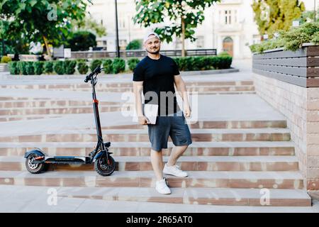 Schöner Mann hält Laptop in der Hand und steht in der Nähe eines Elektrorollers. Aktiver Lebensstil, Rucksacktourist. Ökologie und saubere ökologische Modus der tr Stockfoto