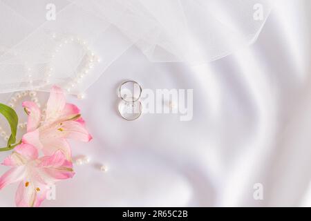 Sanfter Hochzeitshintergrund. Zwei Eheringe aus Platin mit Diamanten auf weißem Satin, Teil des Schleiers der Braut, rosa Blumen Stockfoto
