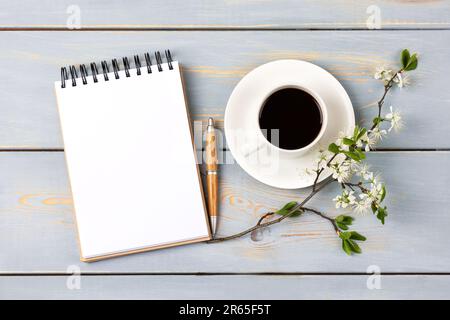 Morgen-Kaffeetasse. Komposition mit geöffnetem leeren Notizblock und Stift, Tasse schwarzen Kaffee, Zweig weißer Kirschblüten auf blauem Holztisch. Festlich Stockfoto