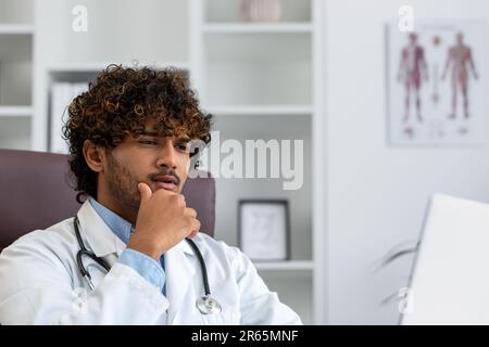 Ernsthaft fokussierter hispanischer Arzt, der in der Arztpraxis arbeitet, Mann in der medizinischen Kittel, der denkt, auf Laptop zu schauen. Stockfoto