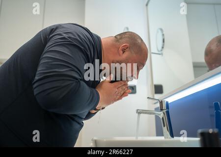 Ein fetter, kahler, bärtiger Mann mittleren Alters wäscht das Gesicht im Badezimmer Haus Internes Waschbecken. Übergewicht und Schweiß Stockfoto