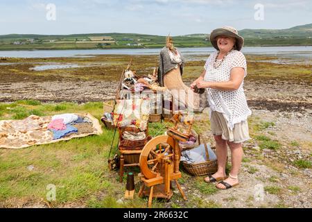 Seniorin mit Drehrad, Wollkugeln und Webartikeln im Freien in Portmagee. Valentia Island im Hintergrund. Stockfoto