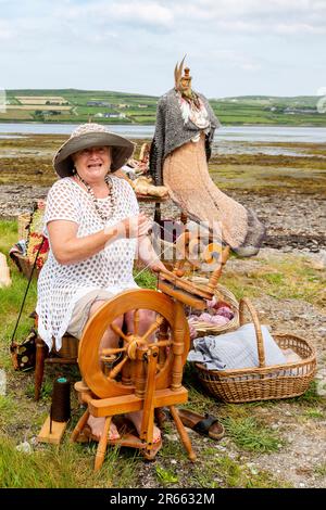 Seniorin mit Drehrad, Wollkugeln und Webartikeln im Freien in Portmagee. Valentia Island im Hintergrund. Stockfoto