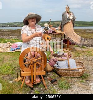 Seniorin mit Drehrad, Wollkugeln und Webartikeln im Freien in Portmagee. Valentia Island im Hintergrund. Stockfoto