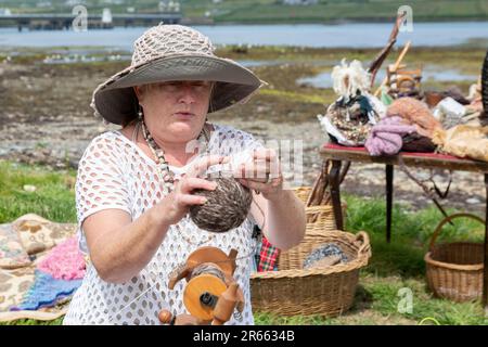 Seniorin mit Drehrad, Wollkugeln und Webartikeln im Freien in Portmagee. Valentia Island im Hintergrund. Stockfoto