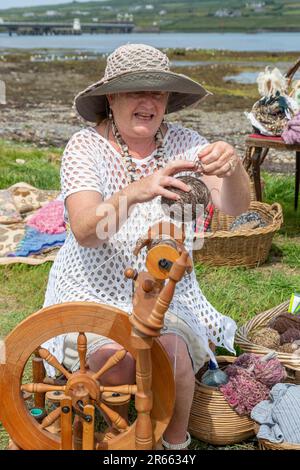 Seniorin mit Drehrad, Wollkugeln und Webartikeln im Freien in Portmagee. Valentia Island im Hintergrund. Stockfoto