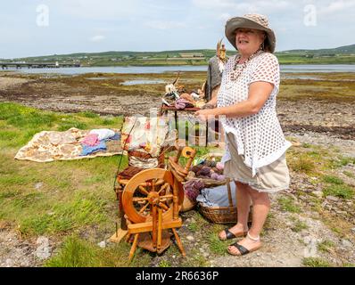 Seniorin mit Drehrad, Wollkugeln und Webartikeln im Freien in Portmagee. Valentia Island im Hintergrund. Stockfoto