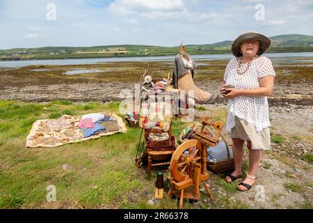 Seniorin mit Drehrad, Wollkugeln und Webartikeln im Freien in Portmagee. Valentia Island im Hintergrund. Stockfoto