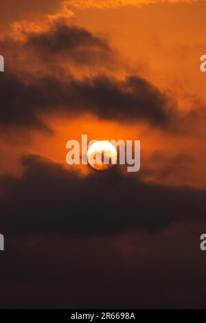 Das intensive orangefarbene Nachglühen des Sonnenuntergangs wirft in dieser beeindruckenden Naturszene einen dramatischen Farbton über die glühenden Wolken und den hellen Mond. Stockfoto