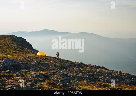 Leute, die Sommerferien in den Bergen verbringen, stehen am Zelt und schauen auf den Sonnenaufgang über dem Tal. Ein aktives Paar wandert in den Bergen. Gelbes Zelt Stockfoto