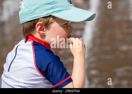 London, Großbritannien - 21. Juni 2023 - Porträt eines weißen kaukasischen Jungen in Mütze, der am Granary Square in Kings Cross Kekse isst Stockfoto