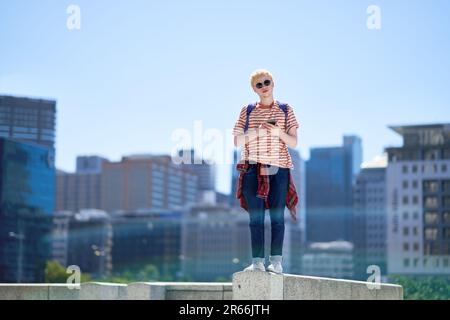 Porträtieren Sie einen jungen Mann mit Smartphone auf dem Sims in der sonnigen Stadt Stockfoto