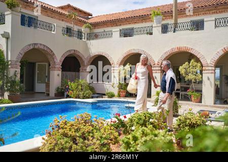 Glückliches Seniorenpaar, das auf der sonnigen Terrasse der Villa am Pool entlang spaziert Stockfoto
