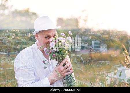 Doppelte Belichtung. Ein gutaussehender Mann schnüffelt Blumen in einem Strauß und schaut in die Kamera. Vor dem Hintergrund einer Stadt, die aus der Höhe genommen wurde. Andere Foc Stockfoto