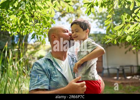 Vater küsst fröhlichen, süßen Sohn mit Down-Syndrom im Sommergarten Stockfoto