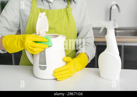 Frau in gelben Handschuhen, die elektrischen Wasserkocher auf einem weißen Tisch reinigt Stockfoto