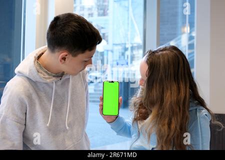 Das Leben von Teenagern am Telefon ein Junge und ein Mädchen sitzen mit ihrem Smartphone an einem Tisch am Fenster und verbringen Zeit im Internet in sozialen Netzwerken, wenn der wi-Fi-Hoodie grau weiß ist Stockfoto