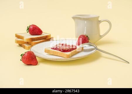 Teller mit leckerem Toast mit Erdbeermarmelade und Milch im Pitcher auf blassgelbem Hintergrund Stockfoto