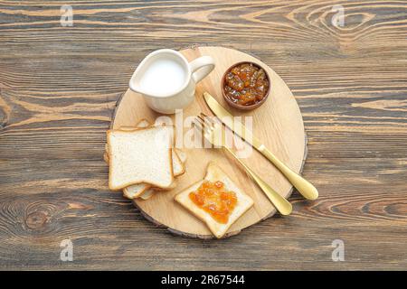 Tafel mit leckeren Toasts mit Aprikosenmarmelade und Milch im Krug auf braunem Holzhintergrund Stockfoto