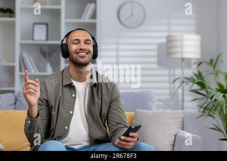 afroamerikaner junger Mann, der zu Hause Musik hört, auf dem Sofa. Er hält ein Telefon und trägt Kopfhörer. Er schloss die Augen und tanzte, erholte sich, genoss. Stockfoto