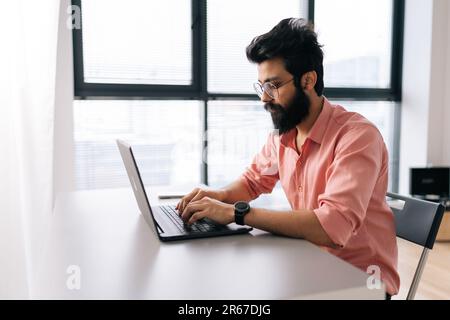 Seitenansicht eines erfolgreichen indischen Programmierers, männlich in Freizeitkleidung, der auf einem Laptop arbeitet, sitzt am Tisch in einem hellen Büro, das am Fenster arbeitet. Stockfoto