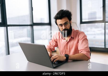 Selbstbewusster indischer Geschäftsmann in lässiger Kleidung sitzt am Schreibtisch mit Laptop und schaut in die Kamera, mit ernstem Ausdruck in einem hellen Büro, das zusammenarbeitet Stockfoto