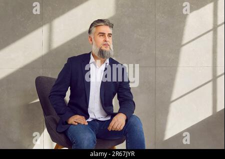 Selbstbewusster, bärtiger, reifer Geschäftsmann, der auf einem Stuhl sitzt, in leerem Raum auf grauem Hintergrund. Stockfoto