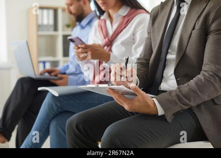 Ein Geschäftsmann, der auf dem Klemmbrett schreibt, während andere im Hintergrund Laptops und Telefone verwenden Stockfoto