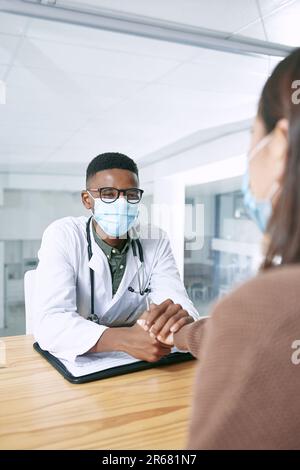 Wie hast du es geschafft? Ein junger Arzt, der bei seiner Patientin sitzt und eine Gesichtsmaske trägt, während er sie in der Klinik tröstet. Stockfoto