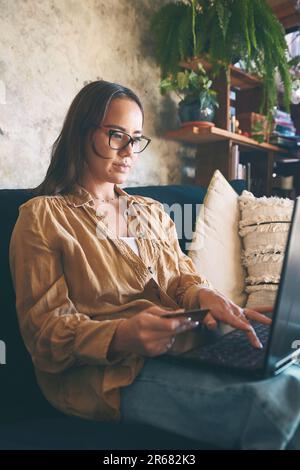 Einkaufen unter Verschluss ist Einkaufen ohne Schlangen. Eine junge Frau, die zu Hause einen Laptop und eine Kreditkarte auf dem Sofa benutzt. Stockfoto