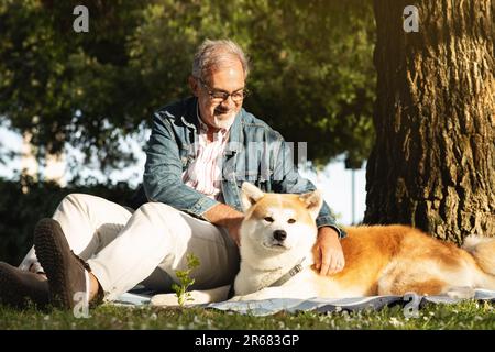 Glücklicher weißer Senior-Mann mit Bart und Brille streichelt den Hund, macht ein Picknick im Park Stockfoto