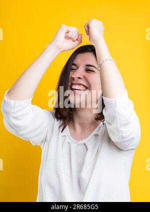 Vor einem gelben Hintergrund hebt eine Frau ihre Arme, tanzt, lacht in einer weißen Bluse und feiert den Erfolg Stockfoto