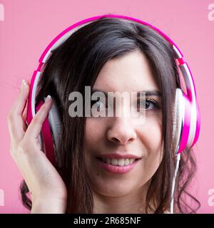 Eine Frau, die sich in Musik und Podcasts vertieft hat, lacht sanft, entspannt. Ihre pinkfarbenen Kopfhörer auf farbenfrohem Hintergrund Stockfoto