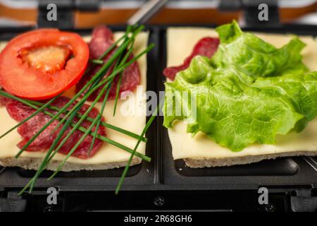 Ein Sandwichmaker mit zwei verschiedenen Sandwiches mit Tomaten, Schinken, Zwiebeln und Käse. Ich bereite eine leichte Mahlzeit zu. Stockfoto