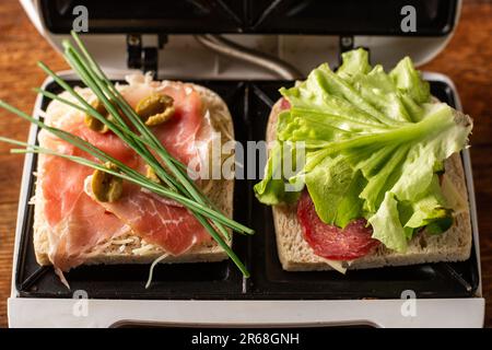 Sandwiches aus Schinken, Gemüse und Käse in einem Sandwichmaker machen. Zwei Toasts mit verschiedenen Füllungen – Nahaufnahme. Stockfoto