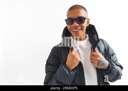 Ein afroamerikanischer Mann in einer Sonnenbrille und einer Pufferjacke, der vor einem weißen Hintergrund vor der Kamera lächelt. Hochwertige 4K-Aufnahmen Stockfoto