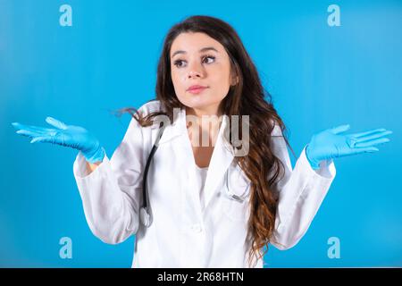 Braunhaariger Arzt in Handschuhen und mit Stethoskop sucht nach Patienten an der blauen Bürowand. Die Krankenschwester sieht verwirrt und besorgt aus Stockfoto