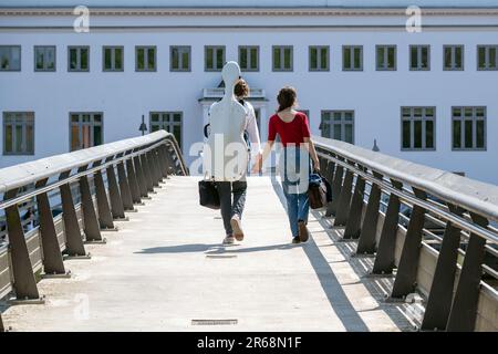 Ein junges Paar Hand in Hand von hinten, das über eine Fußgängerbrücke geht, trägt eine Cello-Box auf dem Rücken, Konzept für den Weg nach vorn mit Liebe an Stockfoto