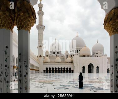 Eine Frau im zentralen Innenhof blickt auf das Minarett und die Kuppeln der Scheich-Zayid-Moschee, Abu Dhabi VAE, die durch den Bogen gesehen werden Stockfoto