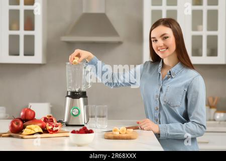 Wunderschöne junge Frau, die Banane in den Mixer gibt, um leckeren Smoothie in der Küche zu erhalten Stockfoto