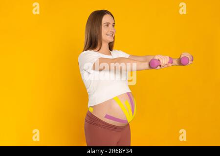 Schwangere Frau mit Kinesio-Bändern auf dem Bauch, die Übungen vor orangefarbenem Hintergrund macht Stockfoto