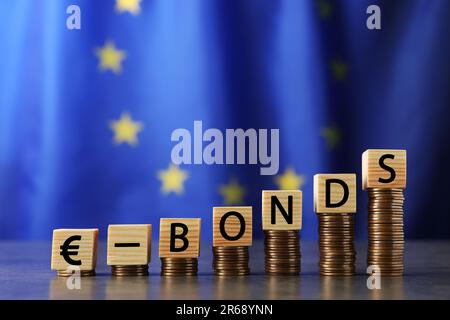 Gestapelte Münzen und Wort-Eurobonds aus Holzwürfeln mit Buchstaben auf einem grauen Tisch gegen die Flagge der Europäischen Union Stockfoto