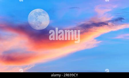 Ein Vollmond erhebt sich in Einem farbenfrohen Sonnenuntergang am blauen Himmel Stockfoto