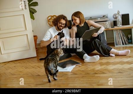 Ein Paar sitzt auf dem Boden und streichelt Katzen, während es Musik auf einem Vinyl-Player mit Schallplatten hört Stockfoto